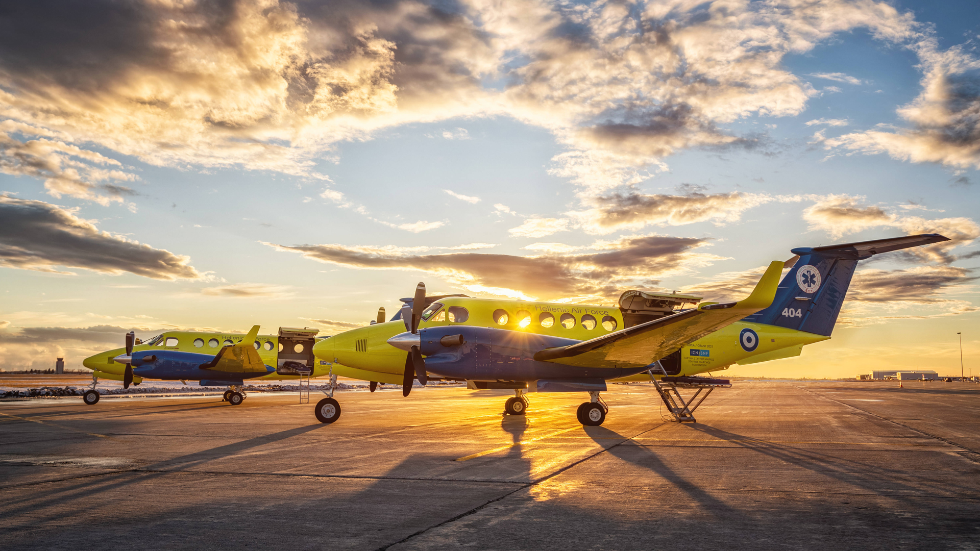 Two yellow and blue medevac propeller planes with the SNF logo sit on the tarmac as the sun sets in the background