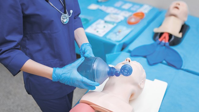 A nurse uses a hand-squeezed breathing assistance device inserted into the mouth of a practice dummy