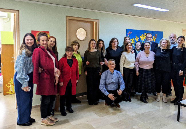 A group of 17 people stand in the corridor of a building, side by side, smiling for the camera.