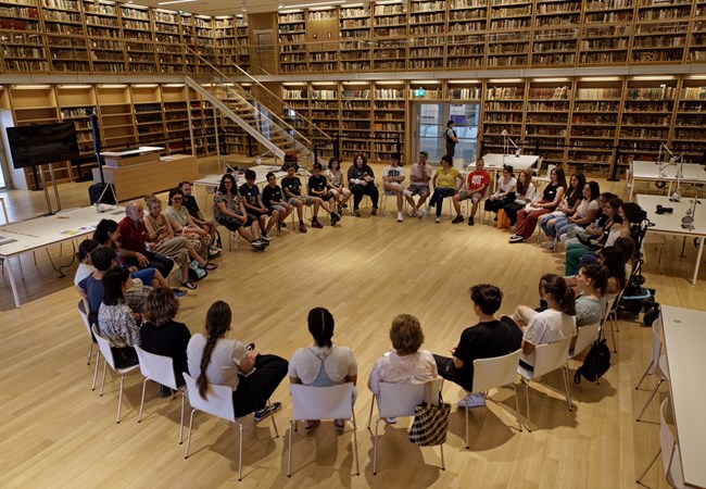 People sit in a large circle of chairs in a warm library context