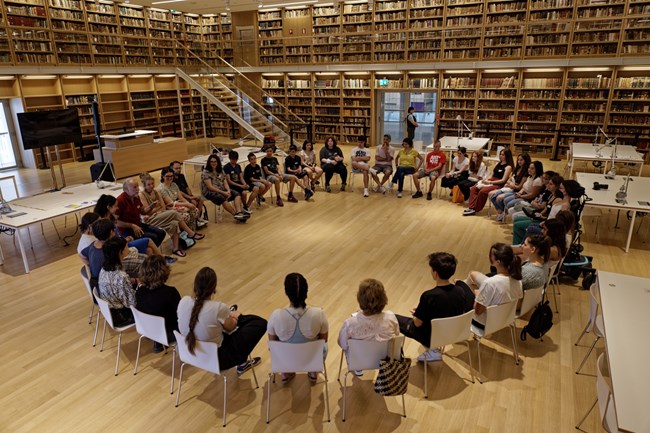 People sit in a large circle of chairs in a warm library context