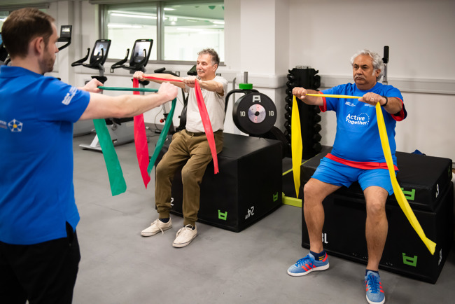 Two seated adults are guided in stretch band exercises by a seated instructor