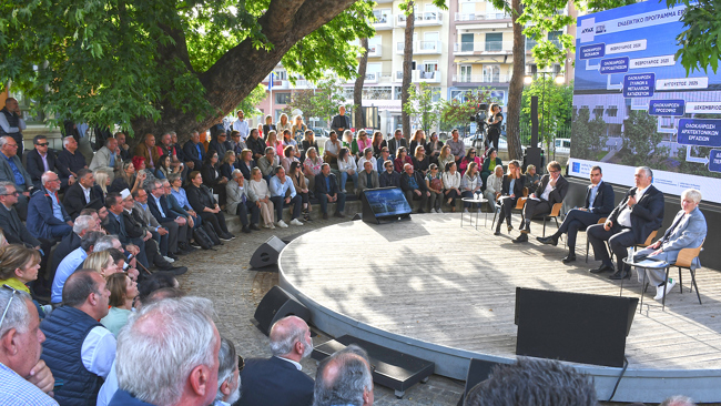 Five people seated on a stage and in front of them there is a diverse seated audience under a tree and they attend the presentation. 