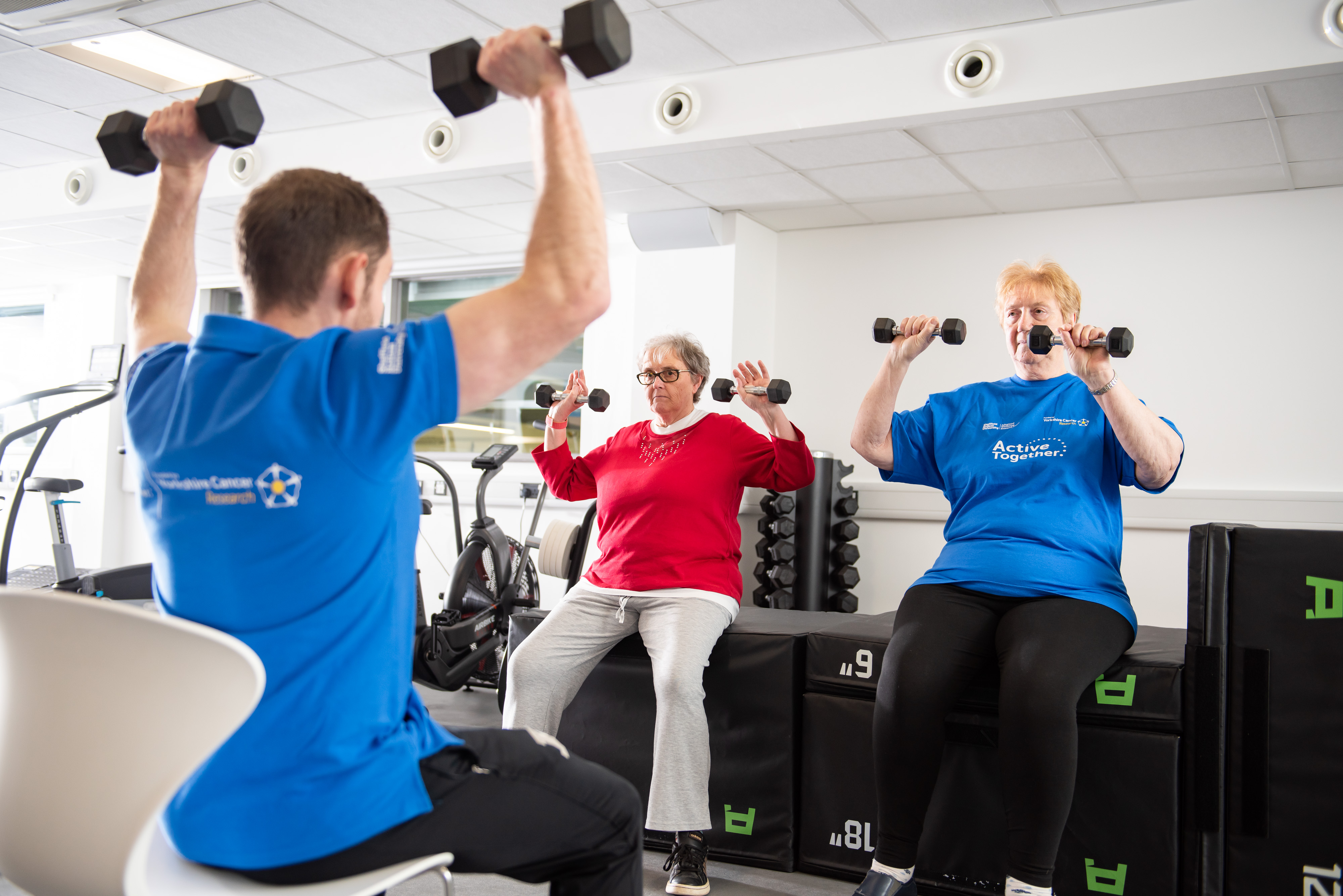 Two seated adults are guided in lifting weights by a seated instructor